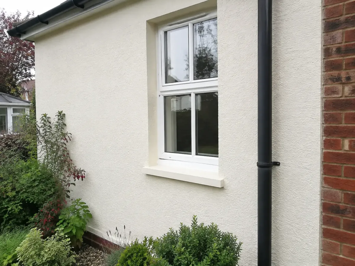 Off-white exterior roughcast wall with a clean window and dark downpipe.